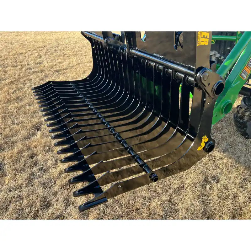 Black metal grapple attachment with evenly spaced curved tines mounted on green tractor loader arm over dry grass field
