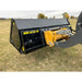 Black hydraulic dump bucket attachment with yellow loader arm pins on dry grass field under cloudy sky