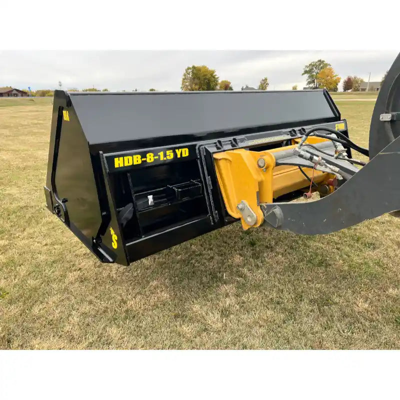 Black steel hydraulic tractor dump bucket attachment mounted on loader arm on grass field with yellow decal