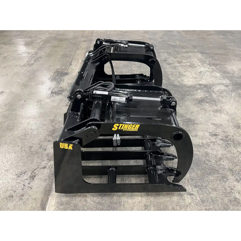 Black metal skid steer grapple attachment with hydraulic cylinders, curved tines, and reinforced frame on concrete floor