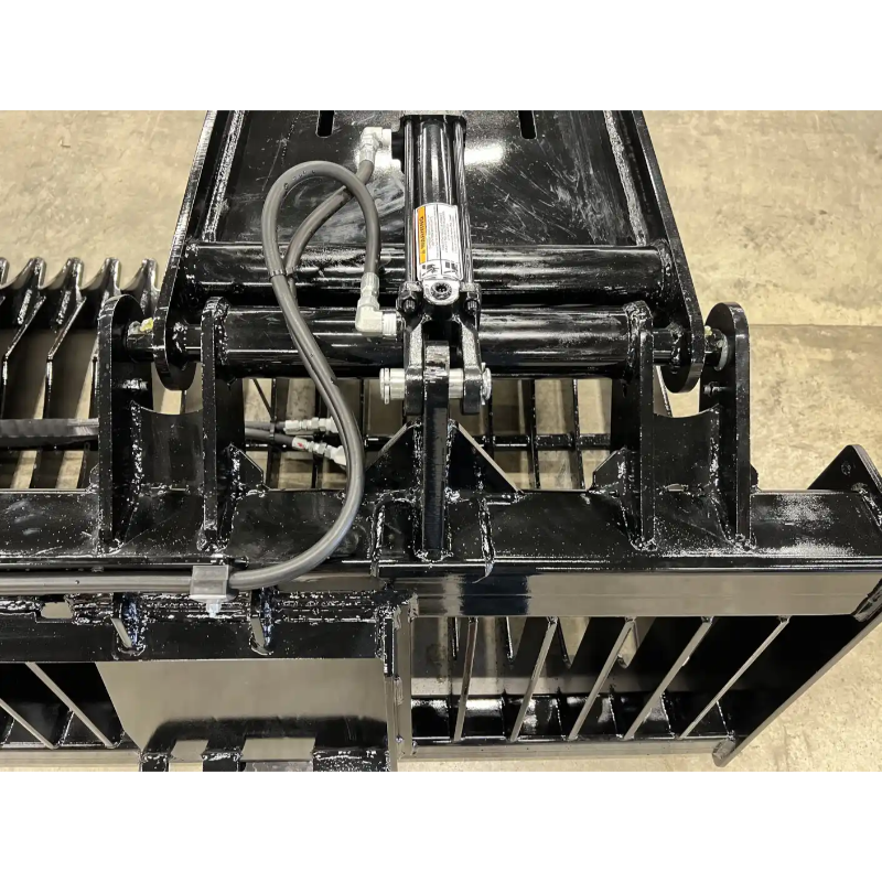 Close-up of black skid steer rock grapple attachment with hydraulic cylinder, hoses, and metal tines on concrete floor