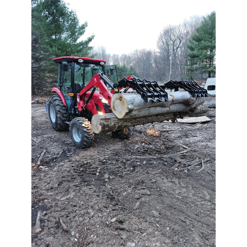 Red compact tractor with enclosed cab lifting multiple large logs using black grapple attachment on dirt ground outdoors