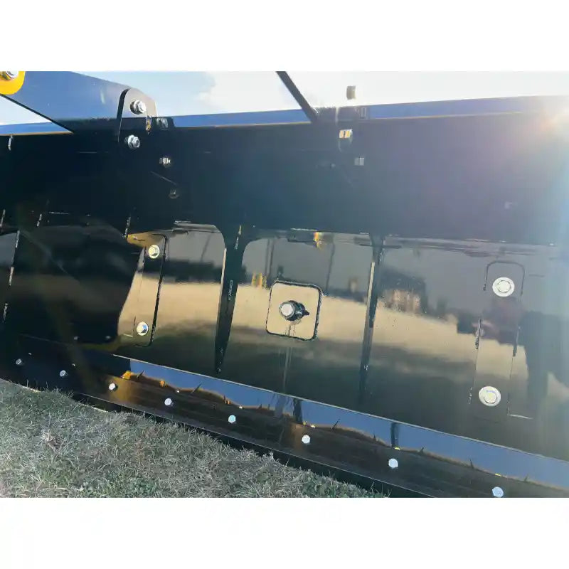 Close-up of black steel bucket attachment with reinforced bottom edge and multiple mounting bolts on grass surface