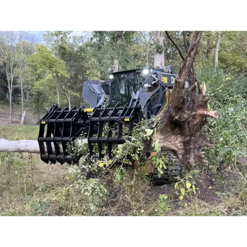 Large tracked skid steer loader with black clam grapple attachment lifting uprooted tree with roots and branches outdoors