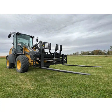 Yellow Deere skid steer loader with black steel pallet fork attachment and extended fork tines on grassy field under cloudy sky