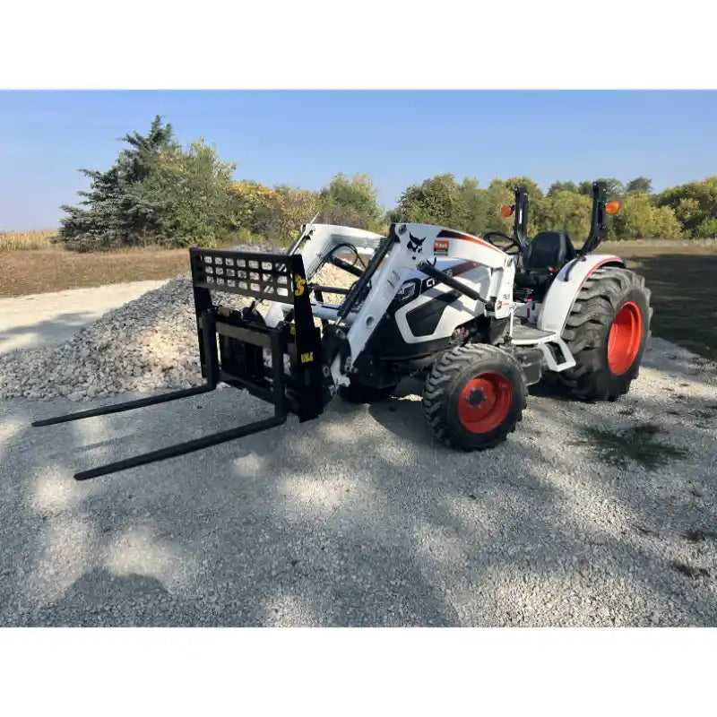 White compact tractor with black steel pallet fork attachment featuring square perforated backrest on gravel outdoors