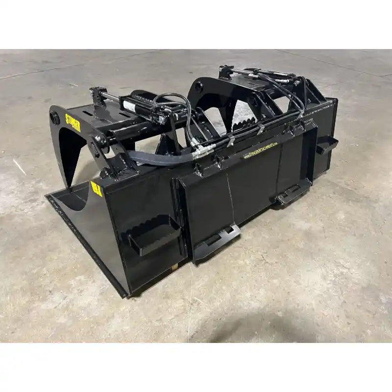 Black steel demolition grapple bucket attachment with hydraulic arms resting on concrete floor in workshop setting
