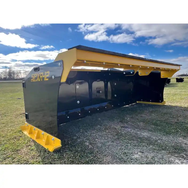 Black and yellow steel snow pusher blade with bolt-on cutting edge resting on grassy field under partly cloudy sky