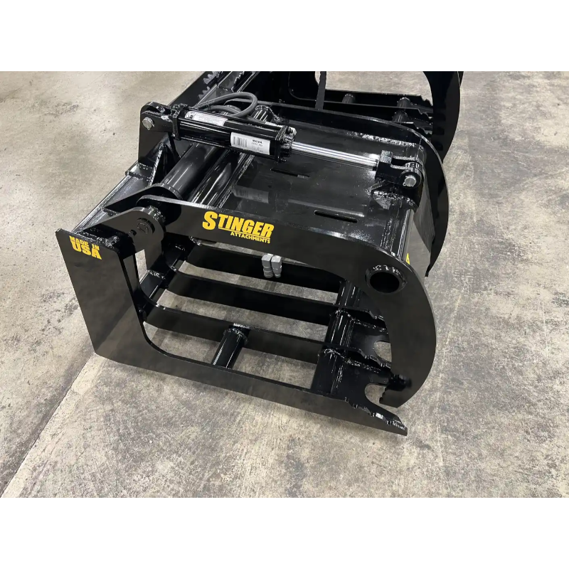 Black skid steer grapple attachment with spaced steel tines, hydraulic cylinder, and welded frame on concrete floor