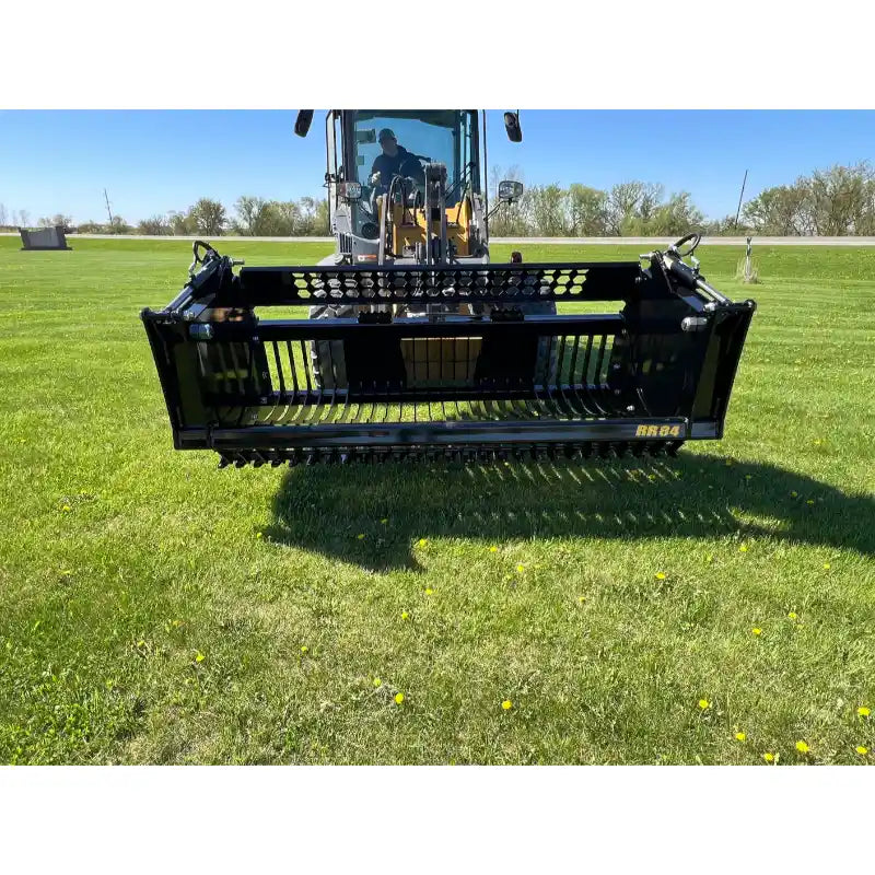 Front view of skid steer mounting a black metal rock bucket reel attachment on green grass under clear sky