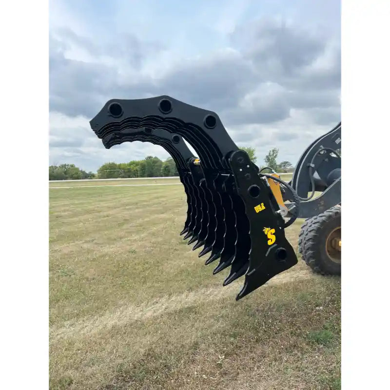 Black multi-tine clam-style grapple attachment mounted on loader arm with hydraulic hoses visible over grassy field