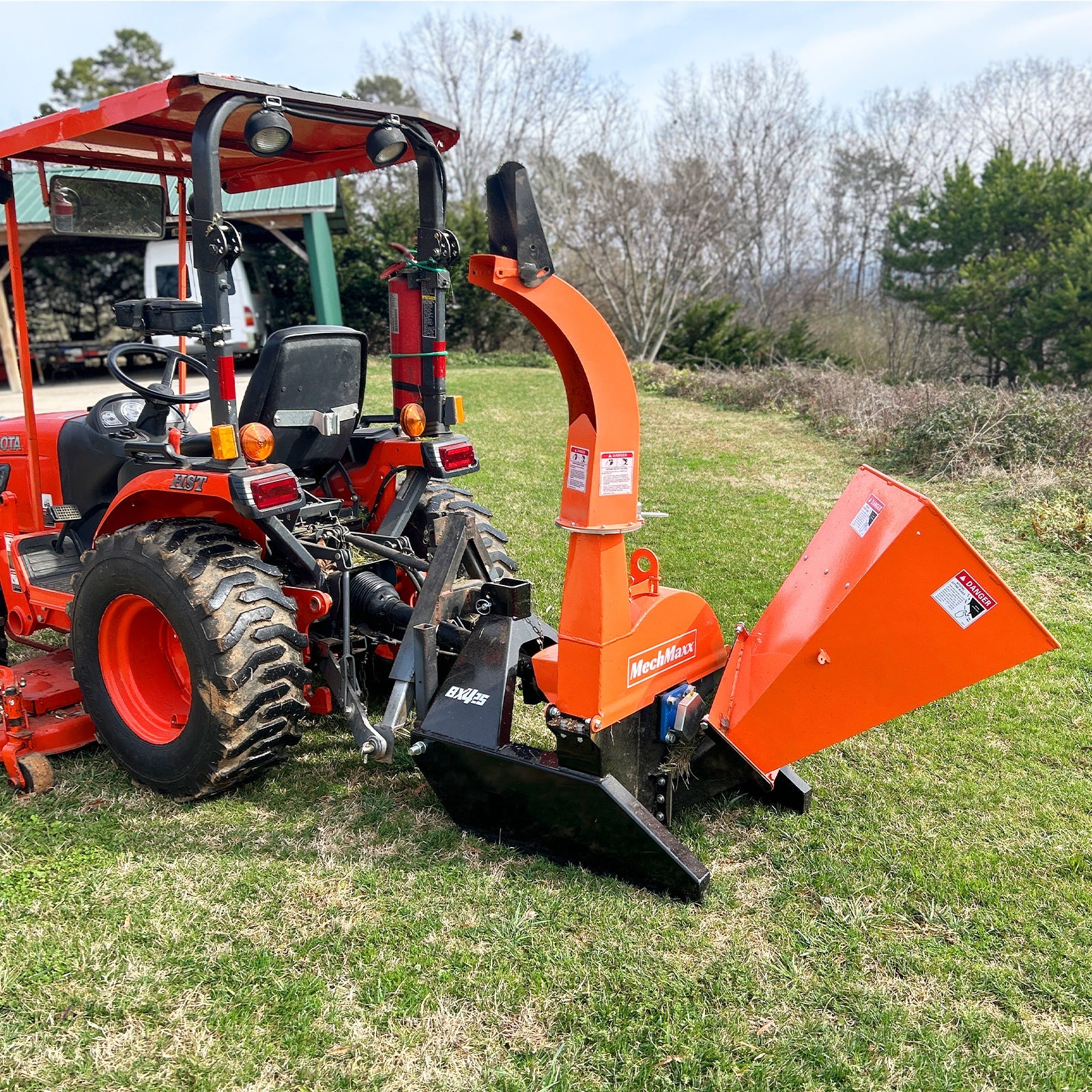 Orange PTO wood chipper attached to tractor with curved discharge chute, angled feed hopper, and black support frame