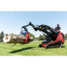 Mini skid steer with red and black hydraulic grapple lifting a large log on a grassy yard with operator wearing sunglasses and hat