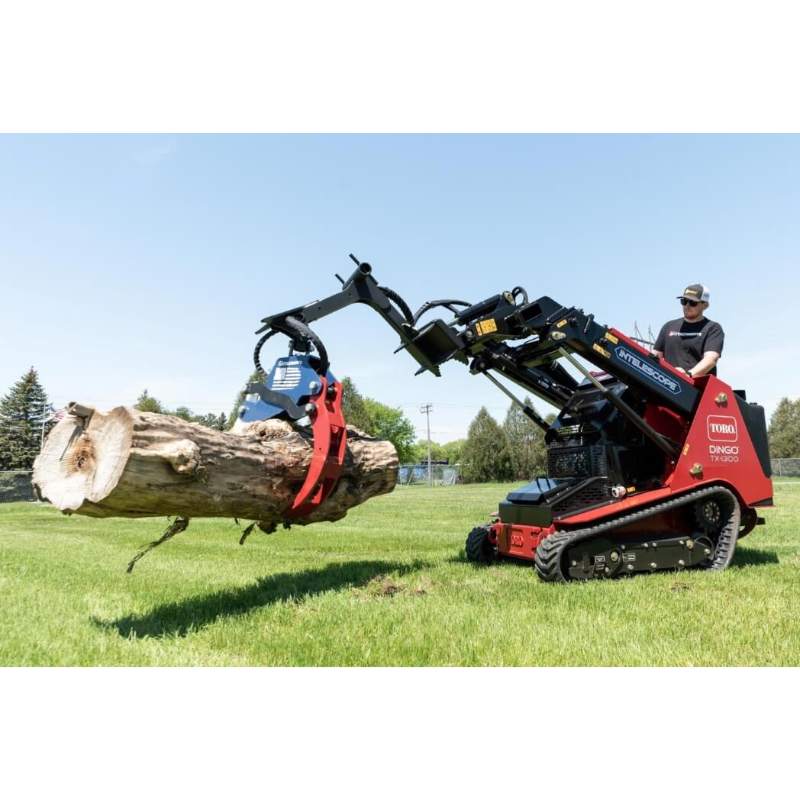 Mini skid steer with red and black hydraulic grapple lifting a large log on a grassy yard with operator wearing sunglasses and hat