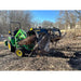 Green tractor with black hydraulic grapple attachment holding large logs in muddy clearing with leafless trees in background