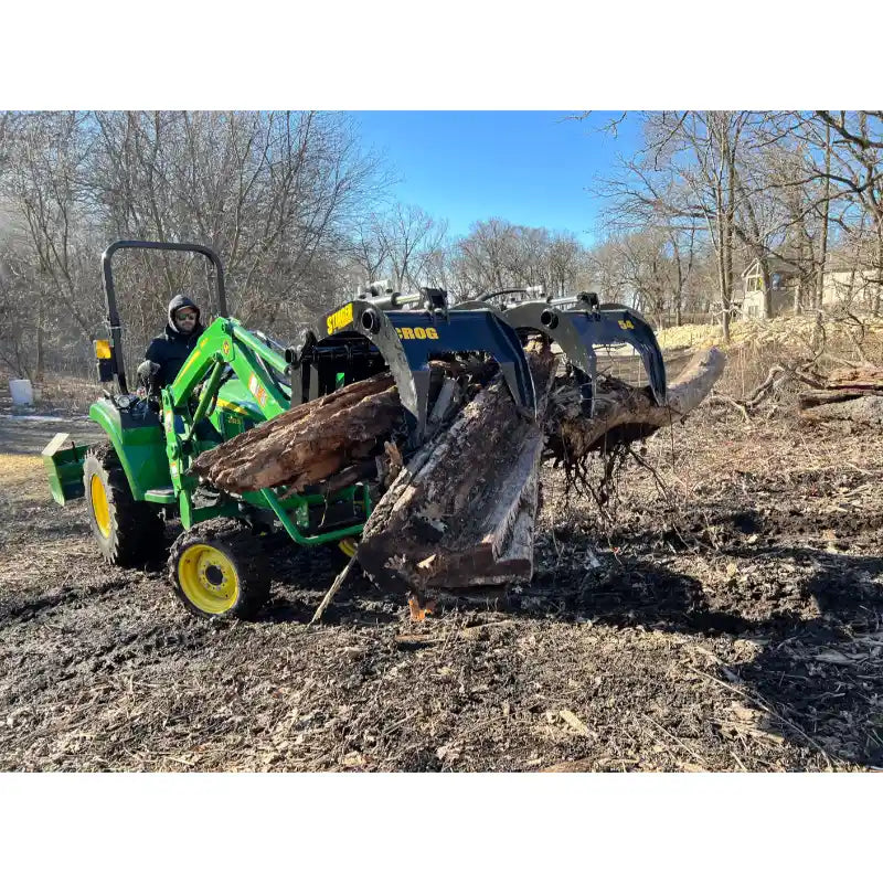 Green tractor with black hydraulic grapple attachment holding large logs in muddy clearing with leafless trees in background