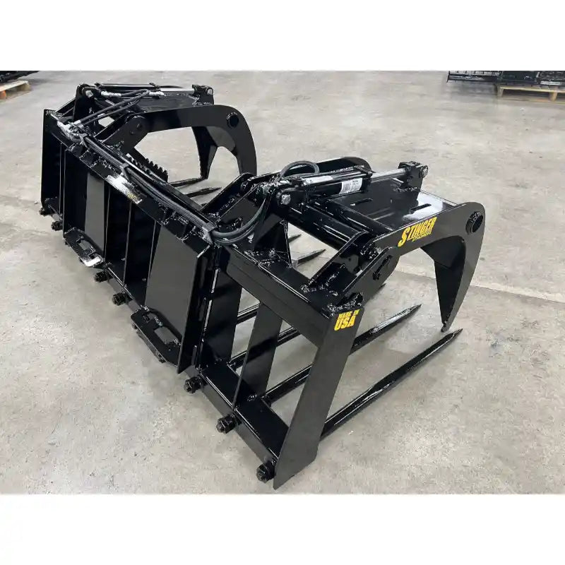 Black-painted metal manure grapple attachment with hydraulic cylinders and wide tines on concrete floor indoors