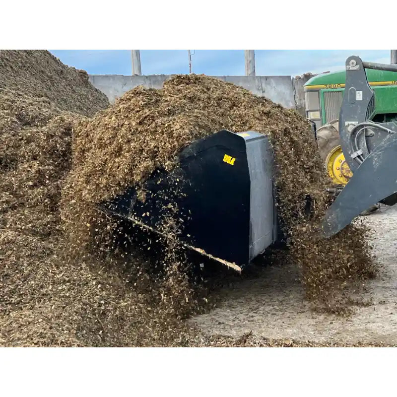 Black steel skid steer high-volume bucket attachment dumping loose mulch at outdoor agricultural site