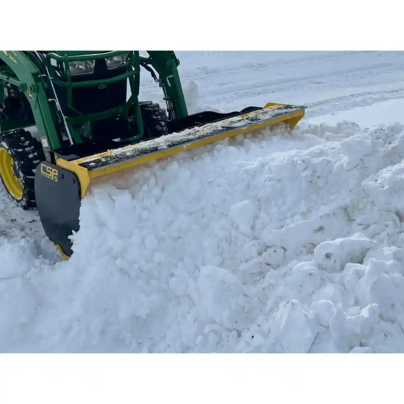 Yellow and black steel snow pusher attachment mounted on green tractor clearing deep snow on flat snowy surface