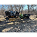 Two black steel rock grapples with curved spaced tines mounted on a green tractor loader arm on dirt ground with leafless trees