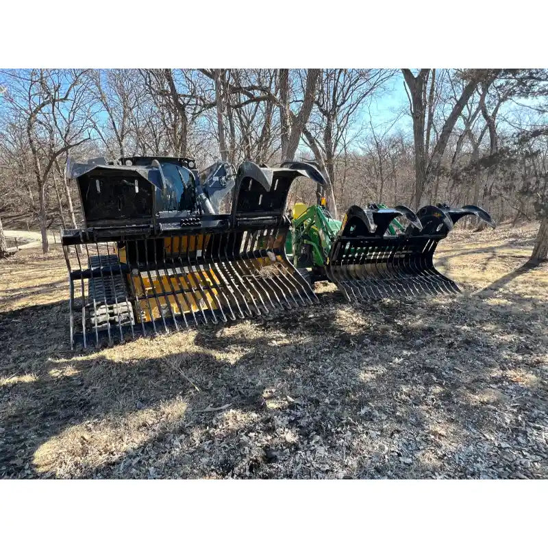 Two black steel rock grapples with curved spaced tines mounted on a green tractor loader arm on dirt ground with leafless trees