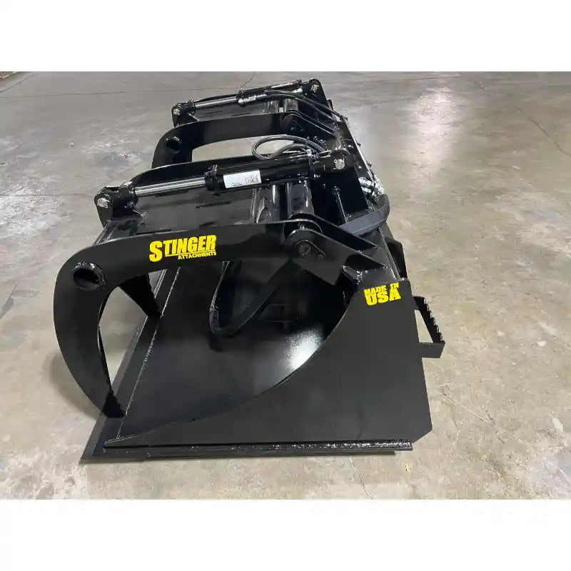 Black steel grapple bucket attachment with hydraulic arms and curved clamp claws on concrete floor indoors