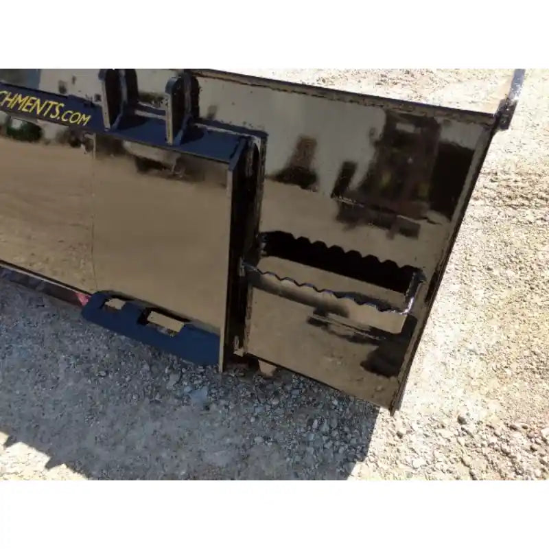 Close-up of black steel skid steer material bucket corner with serrated edge and mounting brackets on gravel surface