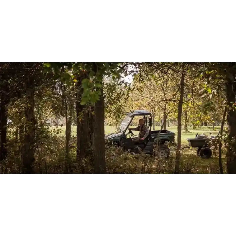 Green utility vehicle towing Swisher 16 cu ft poly cart loaded with materials in wooded outdoor setting, gallery position 1