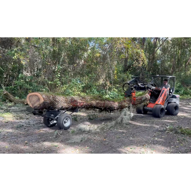 Orange compact loader carrying a large log attached to a black log dolly with wide pneumatic tires on dirt path