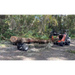 Orange compact loader carrying a large log attached to a black log dolly with wide pneumatic tires on dirt path