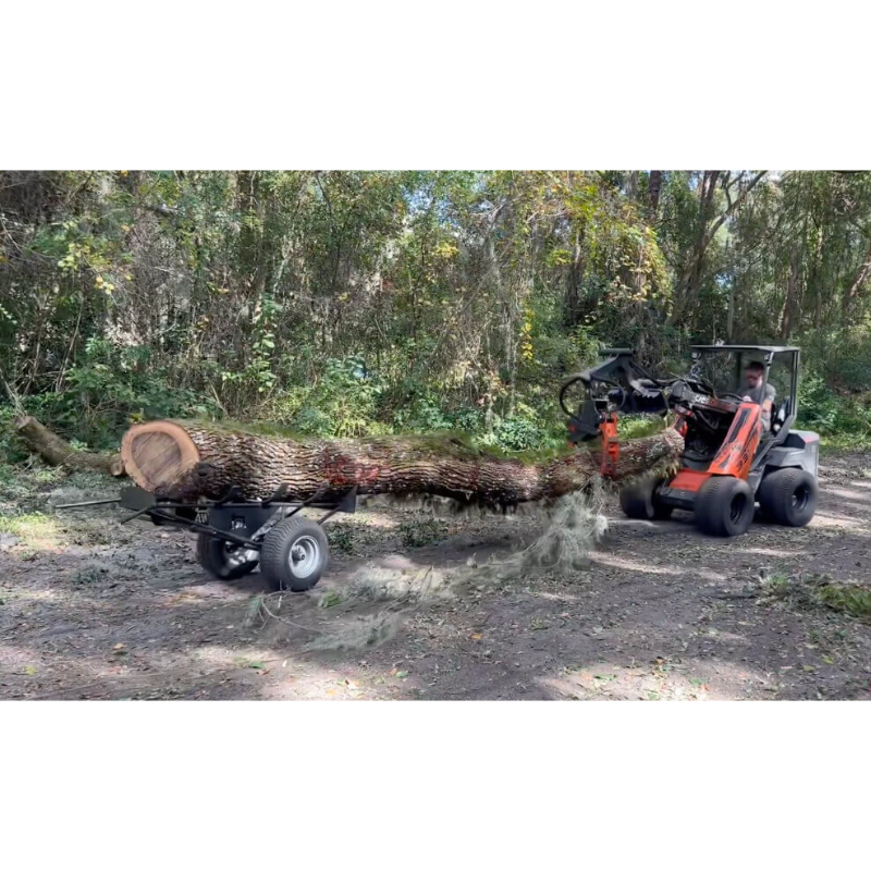 Orange compact loader carrying a large log attached to a black log dolly with wide pneumatic tires on dirt path