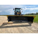 Front view of a skid steer with a black and yellow wide snow pusher blade on a gravel and grass site under blue sky