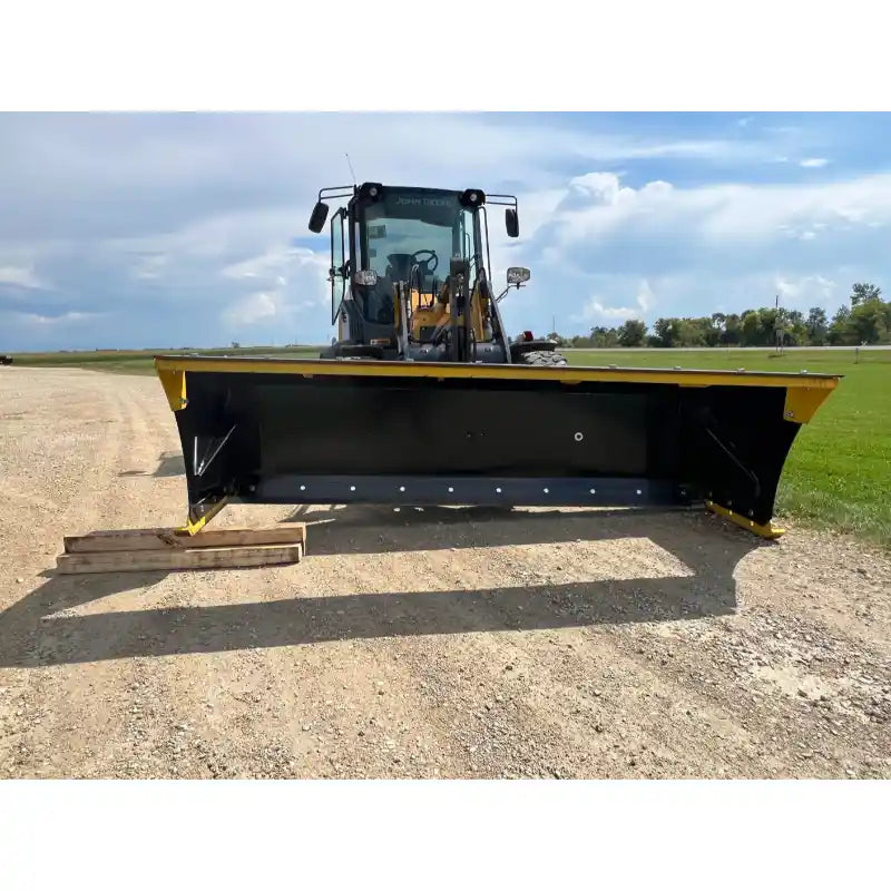 Front view of a skid steer with a black and yellow wide snow pusher blade on a gravel and grass site under blue sky