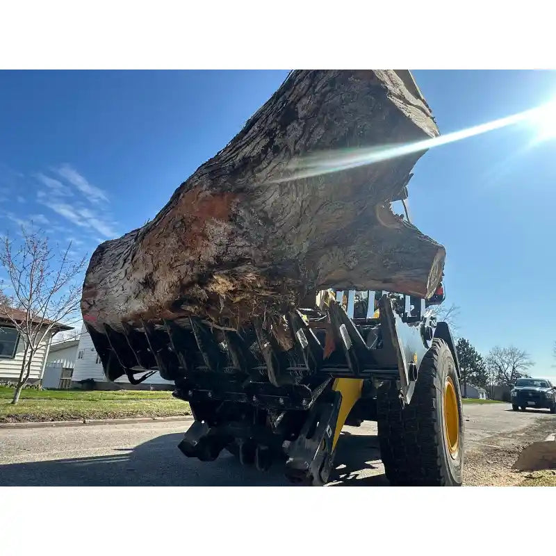 Yellow loader fitted with black hydraulic clam-style grapple lifting a large tree trunk on paved street