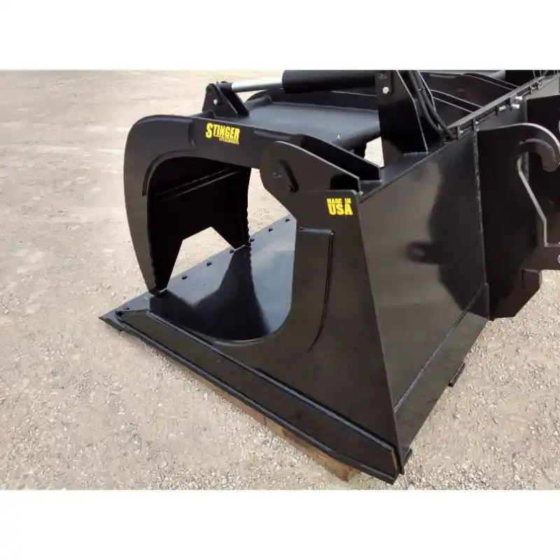 Black steel demolition grapple bucket attachment with hydraulic clamping arms resting on gravel surface outdoors