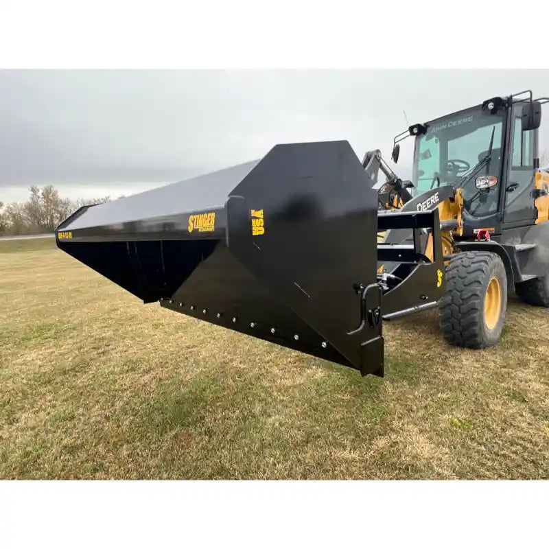 Black steel hydraulic dump bucket attachment mounted on yellow loader with visible cutting edge on grassy field under overcast sky