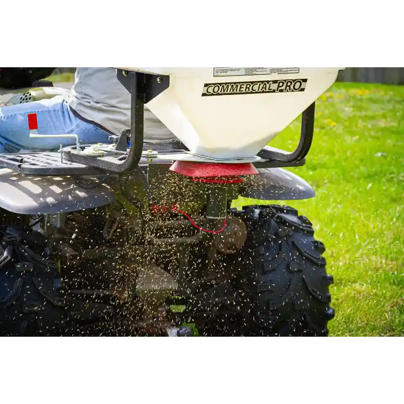 Close-up of Swisher ATV Commercial Pro Spreader mounting and material dispersing beneath ATV on grass, gallery position 1