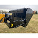 Black steel hydraulic dump bucket attachment with yellow logos mounted on tractor loader arm over grass field under cloudy sky