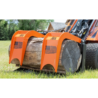 Orange steel grapple attachment with American flag decals gripping a large weathered log on a compact loader outdoors