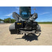 Black skid steer stump grinder attachment with rotating cutting teeth, hydraulic hoses, and protective steel side guard on gravel surface