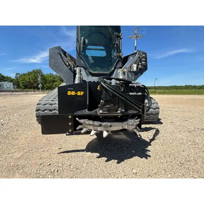 Black skid steer stump grinder attachment with rotating cutting teeth, hydraulic hoses, and protective steel side guard on gravel surface