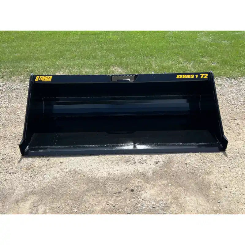 Black steel skid steer material bucket with smooth interior and yellow Stinger Series 1 72 lettering outdoors on gravel and grass