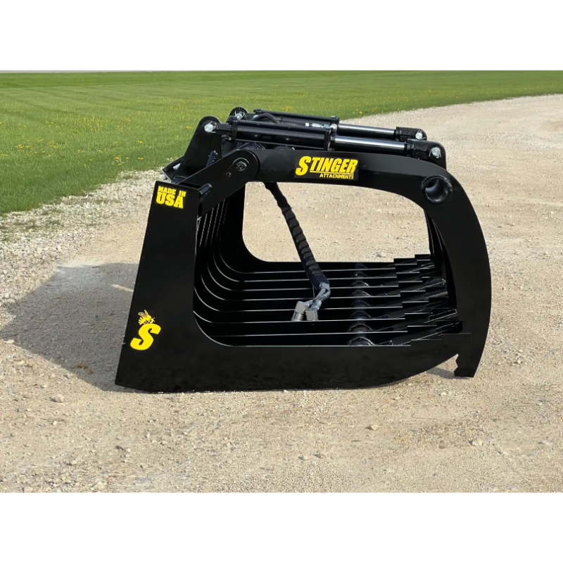 Black steel skid steer root grapple attachment with multiple curved tines and hydraulic cylinders on gravel and grass background
