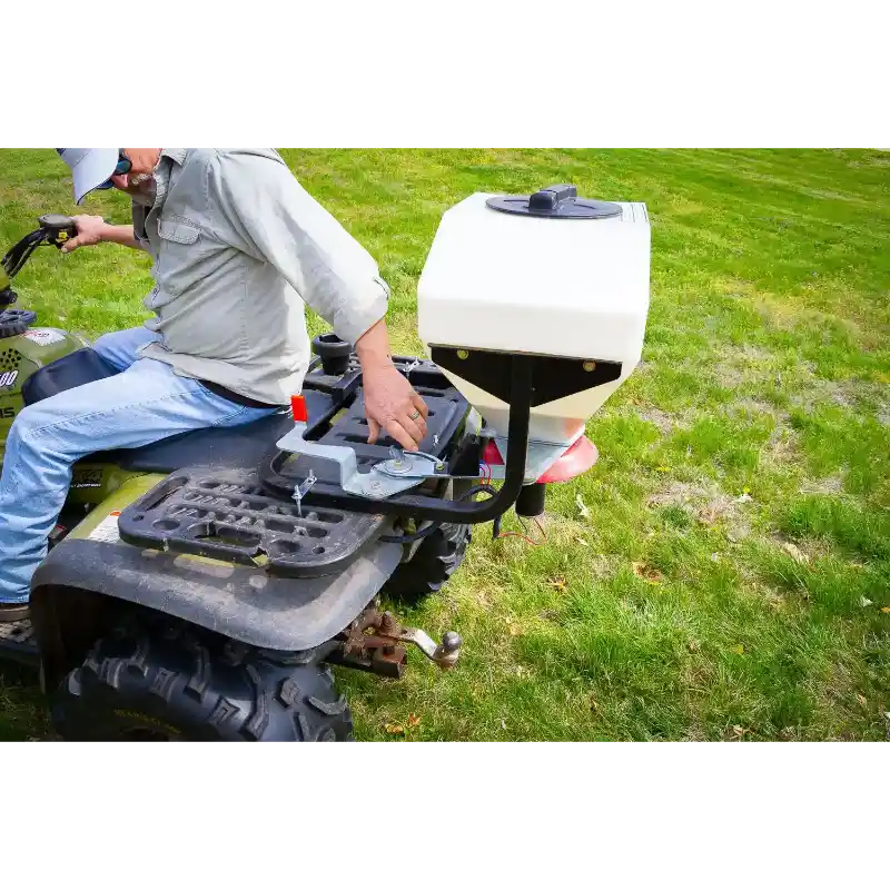 White hopper spreader mounted on rear rack of green ATV with operator adjusting controls, gallery position 1