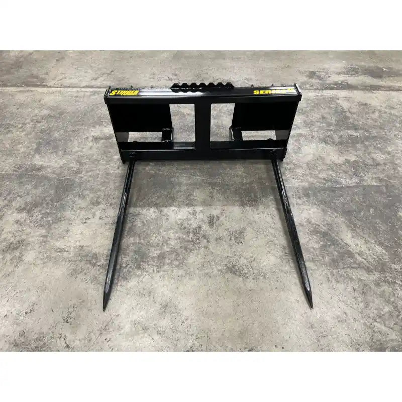 Black steel bale spear attachment with two pointed horizontal tines on concrete floor viewed from front angle