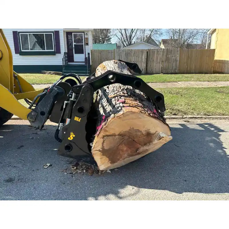Yellow loader equipped with black hydraulic clam grapple attachment holding large cut tree trunk on residential pavement