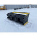 Black and yellow skid steer snow pusher attachment standing on snow-covered ground near industrial buildings outdoors