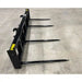 Black metal skid steer attachment with four long steel fork tines on concrete floor with yellow label