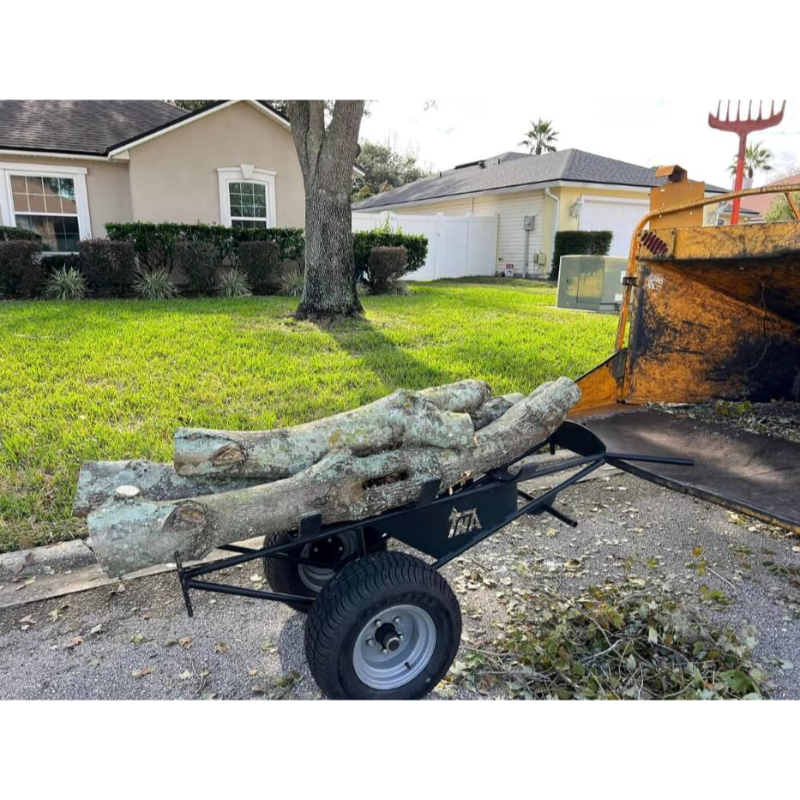 Black steel log dolly with turf tires loaded with cut tree logs beside a yellow wood chipper on gravel driveway