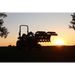 Silhouetted tractor with front loader attachment holding a large metal implement against sunset sky outdoors